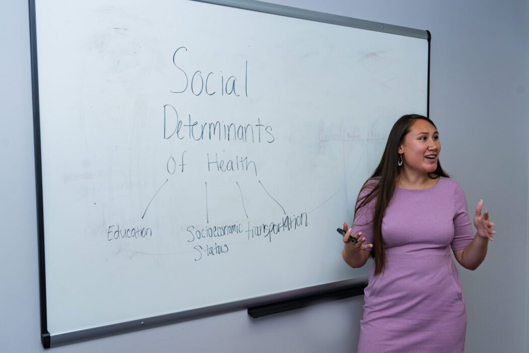 Woman standing in front a whiteboard teaching about the social determinants of health.